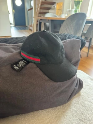 Svart keps med en fin mönster  - Säljer en svart keps  med det klassiska GG-mönstret över hela kepsen. På sidan finns en grön och röd rand som ger en ikonisk touch. Kepsen har böjd skärm och justerbart spänne bak i läder. Perfekt för dig som vill ha en stilren accessoar.