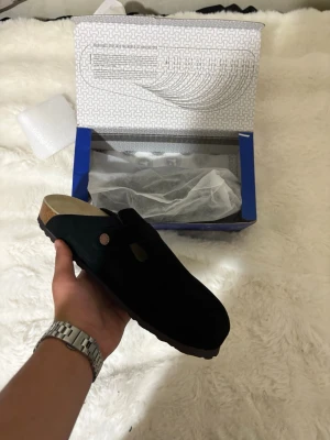 Svarta Birkenstock Boston tofflor - Säljer ett par svarta tofflor i mocka med klassisk korksula och räfflad gummiyta undertill. Tofflorna har en enkel design med en knappdetalj på sidan och öppen häl. Perfekta för chill dagar hemma eller när du vill ha något bekvämt på fötterna.