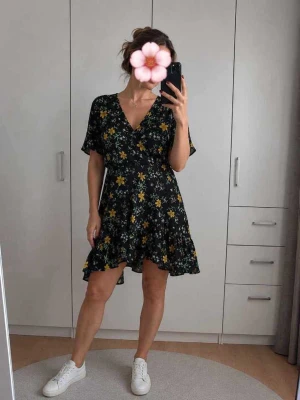 Svart blommig kortklänning med volang - Söt svart kortklänning med gult och grönt blommönster. Klänningen har v-ringning, korta ärmar och volangkant nedtill. Perfekt för sommarens varma dagar och har en avslappnad passform.