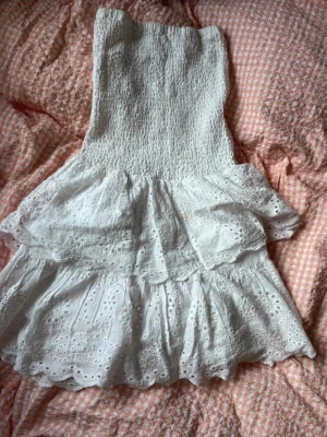 Vit kort klänning med volanger - Söt vit kort klänning med smockad överdel och volangig kjol i broderad bomull. Klänningen är ärmlös och har ett romantiskt uttryck med fina broderade detaljer. Perfekt för varma dagar och somriga tillfällen. En liten fläck längre ner på klänningen men det är inget som märks.