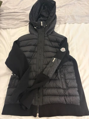 Svart jacka med huva från Moncler - STORLEK L MEN PASSAR M. Säljer en svart pufferjacka från Moncler med huva och dragkedja framtill. Jackan har quiltad design, ribbade stickade ärmar och sidor samt två fickor med knappstängning. Perfekt för kyliga dagar och har en snygg Moncler-logga på ärmen.