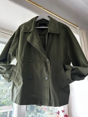 Olivgrön trenchcoat från Monki - Kort trenchcoat i olivgrönt från monki! Den är verkligen så söt men tyvärr så passar den helt enkelt inte så bra på mig.😇