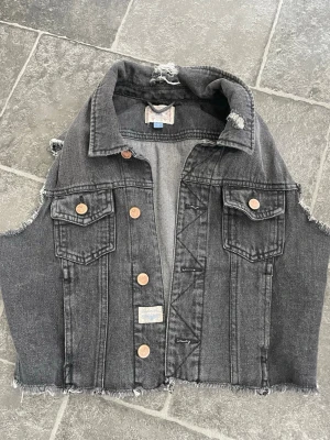 Grå jeansväst med dödskalletryck - Cool grå jeansväst utan ärmar med råa kanter och slitningar. Västen har två bröstfickor med knappar och en stor patch på ryggen med dödskalle, ormar och motorcykelmotiv i svart, vitt, rött och grönt. Perfekt för en edgy streetstil.