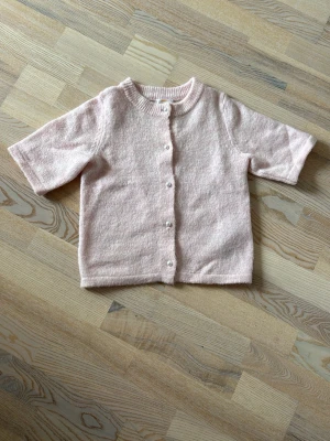 Ljusrosa kofta från Gina Tricot - Supersöt ljusrosa kofta från Gina Tricot med korta ärmar och rund hals. Koftan har knappar framtill och är stickad i ett mjukt material som känns skönt mot huden. Perfekt att slänga över en topp för en söt och enkel look.