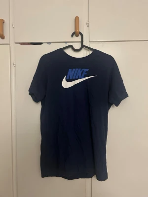 Mörkblå Nike t-shirt med logga - Snygg mörkblå t-shirt från Nike med stor vit och blå logga på bröstet. Klassisk passform och rund halsringning. Tillverkad i mjuk bomull som känns skön mot huden. Perfekt för dig som gillar sportig och enkel stil.