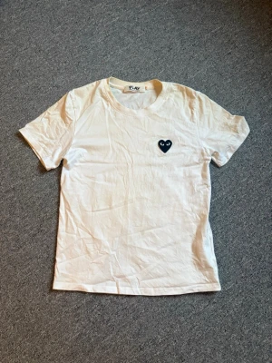 Vit t-shirt Comme des Garçons Play - Vit t-shirt från Comme des Garçons Play med det ikoniska svarta hjärtat med ögon broderat på bröstet. Klassisk rund hals och korta ärmar. Perfekt basic med en twist till din garderob. ( Men i naken och runt kragen så har det blivit gult) 