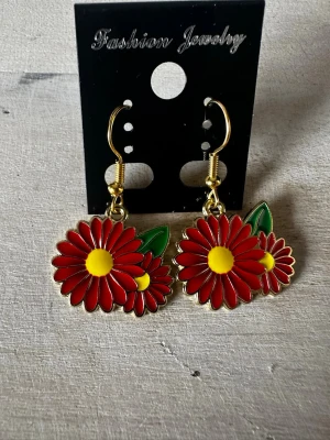 Röda blomörhängen med gul detalj - Unika örhängen i form av röda blommor med gula mittpunkter och gröna blad. Hängena har guldfärgade krokar och en färgglad, lekfull design som verkligen sticker ut. Perfekta för dig som vill addera en färgklick till din stil.