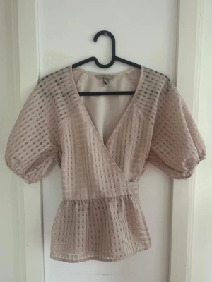 Beige omlottopp från H&M - Säljer en beige omlottopp från H&M med puffiga korta ärmar och v-ringning. Toppen har ett rutigt transparent mönster och markerad midja som ger en snygg siluett. Perfekt för dig som gillar trendiga detaljer och en feminin look.