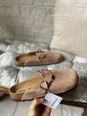 Beige mockasandaler från Birkenstock - Säljer ett par snygga beige sandaler från Birkenstock i mjuk mocka. Modellen har en bred rem med metallspänne och öppen häl. Fotbädden är ergonomiskt utformad och sulan är i kork och gummi för extra komfort. Og Kartong ingår 