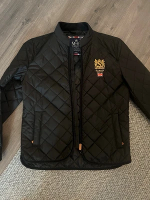 Svart quiltad jacka från Morris - Snygg svart quiltad jacka från Morris med guldbroderad logga och brittisk flagga på bröstet. Jackan har dragkedja framtill, två fickor och en klassisk krage. Perfekt för dig som vill ha en stilren och tidlös look.