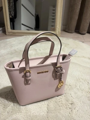 Ljusrosa handväska Michael Kors - Säljer en äkta Michael Kors Jet Set Travel Tote i en jättefin ljusrosa färg med guldfärgade detaljer. 💗  ✨ Modell: Michael Kors Jet Set Travel ✨ Modellnummer: 35T9GTVT0L ✨ Färg: Ljusrosa ✨ Detaljer: Guldfärgade metalldetaljer ✨ Axelrem: Avtagbar och justerbar ✨ Skick: Helt oanvänd – i nyskick!  Väskan är endast förvarad hemma och har aldrig använts. Inga märken, fläckar eller slitage. Perfekt som vardagsväska då den är både stilren och rymlig.  Säljer eftersom den inte kommer till användning. 💕
