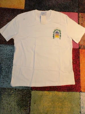 Vit t-shirt med tryck - Vit t-shirt med ett färgglatt tryck på bröstet som föreställer ett motiv med blad och byggnad. Klassisk passform och rund hals. Perfekt för dig som gillar unika prints och enkel stil.