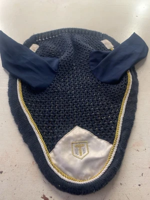Marinblått hästhuvudskydd med brodyr - Säljer ett marinblått hästhuvudskydd med stickad mesh och tygöron. Kantad med vit och gul snodd samt en vit satinpatch med broderad logga framtill. Perfekt för att skydda hästens öron mot insekter och ljud.