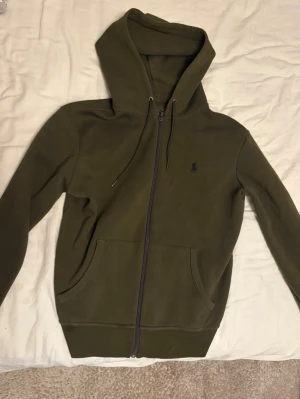 Olivgrön Ralph lauren hoodie med dragkedja - Snygg olivgrön hoodie med dragkedja framtill och två stora fickor. Har huva med dragsko och en liten mörk logga broderad på bröstet. Tillverkad i mjukt sweatshirtmaterial, perfekt för en avslappnad stil.