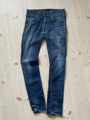 Blå slim fit jeans från Lee - Snygga blå slim fit jeans från Lee med klassisk femficksdesign och lätt slitna detaljer på låren och knäna. Jeansen har normal midja, dragkedjegylf och Lee-lapp bak i midjan. Perfekta för en avslappnad och trendig look.