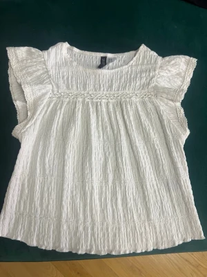 Vit ärmlös blus med volangdetaljer - Söt vit blus i krinklat tyg med volangdetaljer vid axlarna och rund halsringning. Perfekt till jeans eller kjol för en fräsch look. 