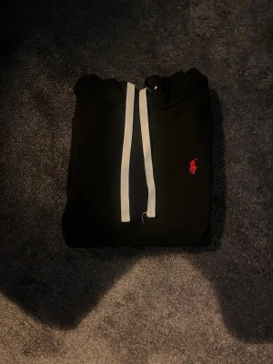 Svart hoodie från Polo Ralph Lauren - Svart hoodie med vit dragsko och klassisk röd Polo Ralph Lauren-logga broderad på bröstet. Hoodien har huva, magficka och ribbade muddar. Perfekt för en chill och stilren look.