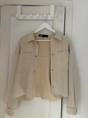 Beige overshirt från Zara - Stilren beige overshirt från Zara med två bröstfickor och klassisk krage. Overshirten har tryckknappar framtill och är tillverkad i ett mjukt, slätt material som ger en avslappnad look. Perfekt att bära öppen eller stängd över en t-shirt. Lite fläckar, men tror de ska gå bort i tvätten