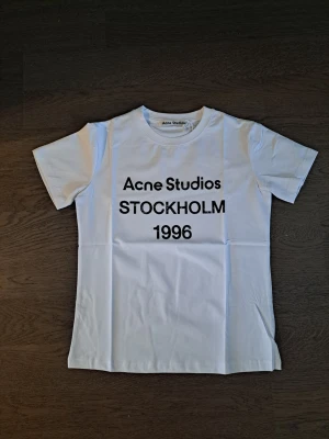 Vit Acne Studios t-shirt med tryck - Säljer en vit t-shirt från Acne Studios med svart texttryck på bröstet där det står 'Acne Studios STOCKHOLM 1996'. Klassisk rak passform, rund hals och korta ärmar. Perfekt till jeans eller shorts för en clean och stilren look.