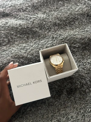 Guldfärgad klocka från Michael Kors - Stilren guldfärgad klocka från Michael Kors med länkarmband och vit urtavla. Klockan har flera små urtavlor och tydliga markeringar, vilket ger en lyxig och modern känsla. Perfekt accessoar för dig som vill sticka ut.