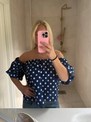 Blå offshoulder topp med vita prickar - Säljer en blå offshoulder topp med vita prickar och puffiga korta ärmar. Toppen har en lös passform och resår vid axlarna för en bekväm look. Perfekt för dig som gillar trendiga och lekfulla plagg.