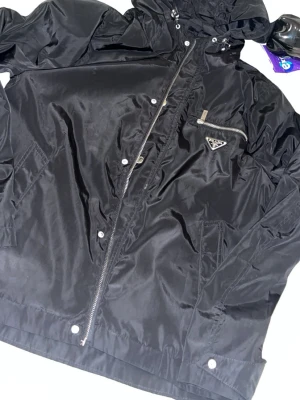 Svart vindjacka  - Snygg svart vindjacka  i glansigt nylonmaterial. Jackan har dragkedja framtill, tryckknappar, huva och en bröstficka med silverfärgad dragkedja i metall. Perfekt för en trendig och sportig look.