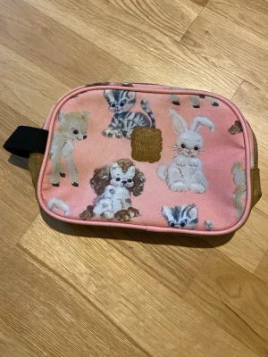 Rosa necessär med djurmotiv - Söt rosa necessär med tryck av olika djur som katt, kanin och hund. Väskan har dragkedja, svart handtag och detaljer i beige. Perfekt för småprylar eller smink. Materialet är tyg med plastdetaljer och motivet är riktigt gulligt.