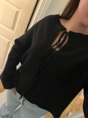 Svart tröja med snörning fram - Snygg svart tröja med långa ärmar och trendig snörning framtill vid halsen. Tröjan har en avslappnad passform och är perfekt att matcha med jeans. Materialet ser mjukt och bekvämt ut, och modellen har en lätt croppad känsla.
