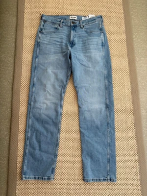 Ljusblå raka jeans Wrangler W32 L32 - Säljer ett par helt oanvända ljusblå jeans från Wrangler med klassisk rak passform och fem fickor. Jeansen har subtila slitningar framtill och tydliga sömmar på bakfickorna. Tillverkade i denim med en skön och avslappnad look. Nyskick 