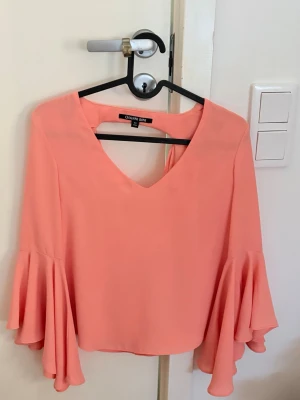 Korallfärgad blus från Gianni Bini - Supersnygg korallfärgad blus från Gianni Bini köpt i USA- med v-ringning och vida, volangiga ärmar. Blusen har en lös passform och är tillverkad i ett lätt, mjukt tyg som ger ett flowy intryck. 