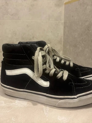 Svarta höga sneakers med vit sula - Klassiska svarta höga sneakers med vit sula och vit sicksackad sidodetalj. Skorna har vita snören och kontrastsömmar, samt vadderad ankel. Materialet är en mix av mocka och textil. Perfekta för en streetwear-look.