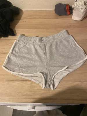 Grå mjukisshorts med vit kant - Säljer ett par ljusgrå mjukisshorts med vit kant och elastisk midja. Shortsen har en sportig look och är perfekta för chill dagar eller träning. Tillverkade i mjukt bomullsmaterial för maximal komfort.