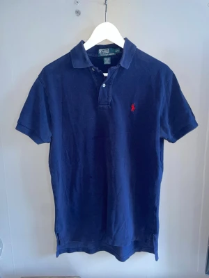 Marinblå pikétröja från Polo Ralph Lauren - Klassisk marinblå pikétröja från Polo Ralph Lauren med korta ärmar och krage. Tröjan har två knappar vid halsen och den ikoniska röda logotypen broderad på bröstet. Tillverkad i mjuk bomull för en bekväm känsla.