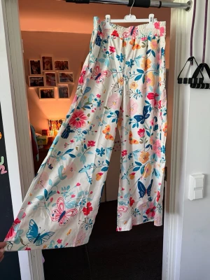 Färgglada byxor med fjärilar - Säljer ett par byxor i vitt tyg med färgglatt blommigt och fjärilsmönster i blått, rosa, orange och turkost. Byxorna har breda ben och resår i midjan för extra komfort. Storlek XL 42 helt nya