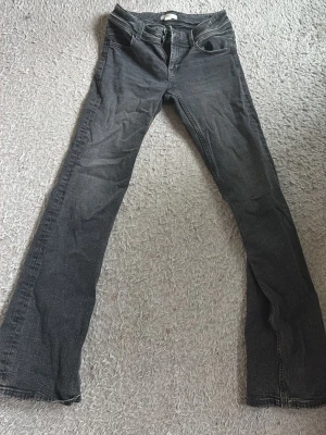 Grå bootcut jeans med låg midja  - Säljer ett par grå/ svart bootcut jeans med hög midja och klassiska fem fickor. Jeansen har en snygg tvättad look och är tillverkade i denim med lite stretch. Perfekta för dig som gillar en retro vibe och vill ha ett par jeans som sticker ut.
