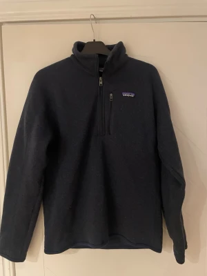 Mörkblå fleece från Patagonia - Mysig mörkblå fleece från Patagonia med halv dragkedja och hög krage. Perfekt för lager-på-lager och outdoor. Klassisk logga på bröstet och mjukt material som håller värmen. Långärmad och enkel att matcha till det mesta.