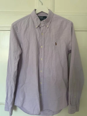 Ljuslila skjorta från Polo Ralph Lauren - Säljer en ljuslila skjorta från Polo Ralph Lauren i slim fit-modell. Skjortan har klassisk krage med knappar, långa ärmar och den ikoniska broderade loggan på bröstet. Tillverkad i bomull och perfekt för en clean och stilren look.
