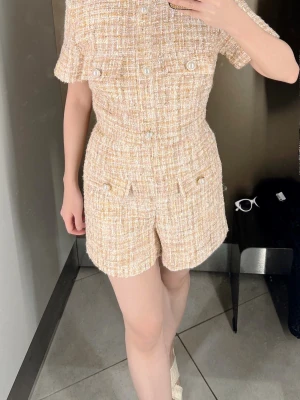 Beige tweed jumpsuit med pärlknappar - Kortärmad jumpsuit i beige tweed med snygga pärlknappar och fickdetaljer fram. Figurnära passform och korta ben ger en chic och trendig look. Perfekt för dig som vill ha en elegant och modern stil.