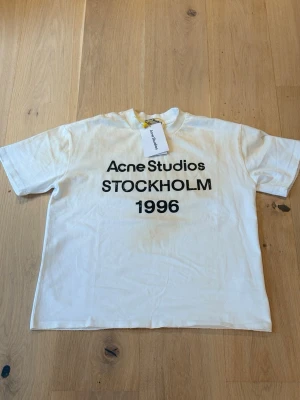 Vit Acne Studios t-shirt  - Vit t-shirt från Acne Studios med svart text 'STOCKHOLM 1996' på bröstet. Klassisk passform med rund hals och korta ärmar. Tillverkad i mjuk bomull, perfekt för en clean och stilren look.          Hör av dig vid minsta fundering😊