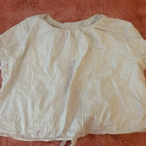Vit randig blus med knyt - Söt vit blus med tunna vertikala ränder och korta ärmar. Toppen har ett knyt framtill som ger en avslappnad vibe och är tillverkad i ett lätt bomullsliknande material. Perfekt till jeans eller kjol för en fräsch look.