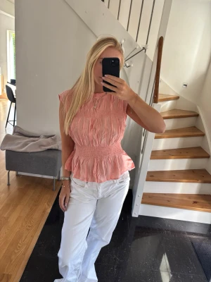 Ljusrosa blus med volangdetaljer - Supersöt ljusrosa blus med volangkant och smock i midjan. Blusen är ärmlös och har en rund halsringning. Materialet ser ut att vara lätt och luftigt, perfekt för varmare dagar. Passformen är normal och toppen ger en feminin siluett.