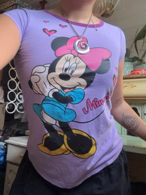 Lila Minnie Mouse t-shirt - Söt lila t-shirt med tryck av Minnie Mouse i färgglada toner och rosa detaljer. T-shirten har korta ärmar och en rund halsringning med lila kant. Perfekt för dig som älskar Disney och vill ha en lekfull stil. Barnstorlek men sitter såhär på mig med storlek 38!Budas ut på live 25 augusti 19.00.