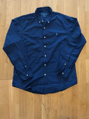 Mörkblå skjorta från Ralph Lauren - Klassisk mörkblå skjorta från Ralph Lauren med button-down krage och vita knappar. Skjortan har lång ärm och den ikoniska broderade loggan på bröstet. Tillverkad i bomull för en skön och stilren look.