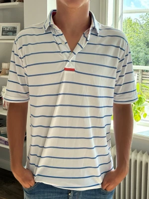 Vit och blårandig pikétröja från Footjoy - Vit pikétröja med blå horisontella ränder och en röd detalj vid knäppningen. På ryggen finns en bred röd rand och ett litet FJ-tryck. Tröjan har krage och korta ärmar, perfekt för en avslappnad stil.