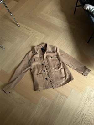 Beige jacka med fickor - Snygg beige jacka från J. Lindeberg med klassisk krage och fyra framfickor. Jackan har svarta knappar framtill och på ärmsluten samt raka sömmar för en clean look. Rak modell med normal passform. Perfekt att slänga över en hoodie eller t-shirt för en avslappnad stil, snygg till jeans och ger en väldigt clean look.