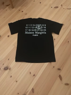 Maison Margiela svart t-shirt - Svart t-shirt från Maison Margiela med ikoniskt tryck på ryggen i vitt och siffror samt logga. Klassisk passform och rund hals. Tillverkad i mjuk bomull som känns skön mot huden. Perfekt för dig som gillar streetwear och vill ha något stilrent men ändå unikt.