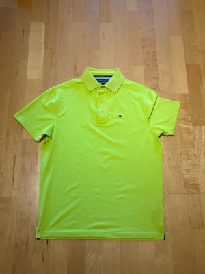 Neongul piké golftröja från Tommy Hilfiger - Snygg neongul pikétröja från Tommy Hilfiger golf med klassisk krage och korta ärmar. Tröjan har knappslå med två knappar och en liten logga på bröstet. Tillverkad i mjukt och lätt material som passar perfekt till sommaren.