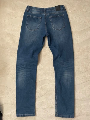 Lagency Jeans - Snygga blå jeans med raka ben och klassiska bakfickor. Jeansen har en lätt tvättad look med subtila slitningar på benen och en svart patch vid linningen. Perfekta för en avslappnad och trendig stil.