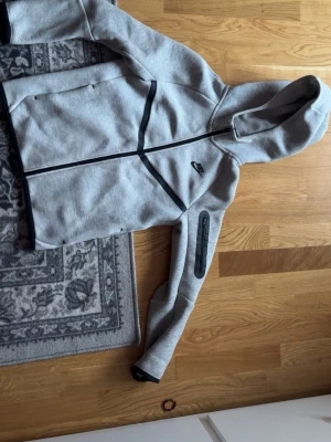 Grå Nike hoodie med svarta detaljer - Snygg grå hoodie från Nike med svarta dragkedjor och detaljer. Hoodien har hel dragkedja framtill, huva och en extra ficka med dragkedja på ena ärmen. Perfekt för en sportig och avslappnad stil.