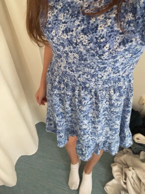 Blå blommig kortklänning - Supersöt blå kortklänning med små vita blommor och korta puffärmar. Klänningen har en avslappnad passform och är perfekt för varma dagar. Tillverkad i ett mjukt och lätt tyg som känns skönt mot huden.